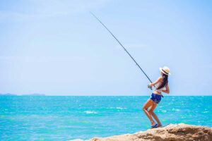 Woman standing on a rocky beach, fishing rod cast over turquoise ocean under a clear blue sky, wearing a sun hat and shorts.