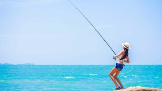 Woman standing on a rocky beach, fishing rod cast over turquoise ocean under a clear blue sky, wearing a sun hat and shorts.