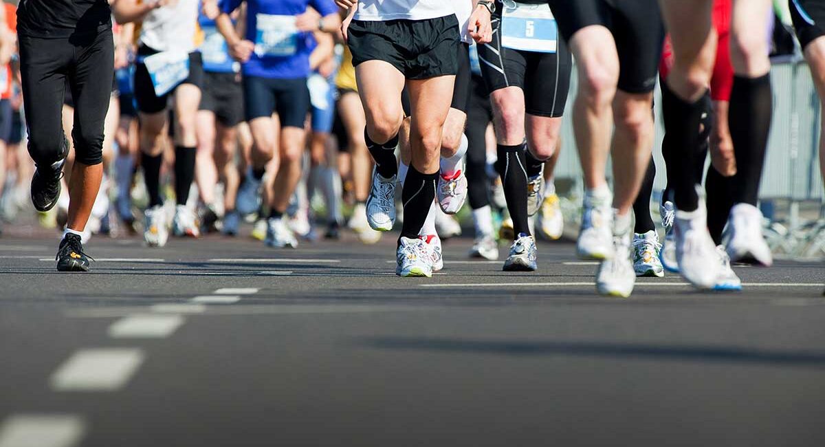Group of runners in colorful athletic gear sprinting on a road, race bibs visible on their chests/waists.