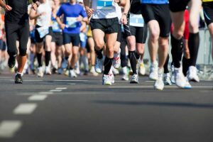 Group of runners in colorful athletic gear sprinting on a road, race bibs visible on their chests/waists.