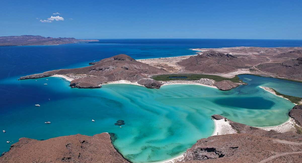 Aerial view of a turquoise coastal lagoon with white sand beaches framed by brown rocky hills and a deep blue sea in the distance.