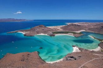 Aerial view of a turquoise coastal lagoon with white sand beaches framed by brown rocky hills and a deep blue sea in the distance.