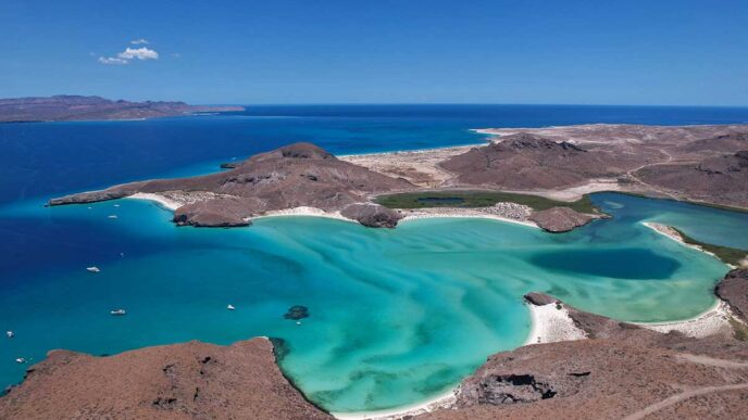 Aerial view of a turquoise coastal lagoon with white sand beaches framed by brown rocky hills and a deep blue sea in the distance.