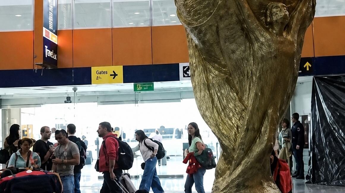 Large gold World Cup trophy sculpture in an airport terminal, with travelers and luggage around it.