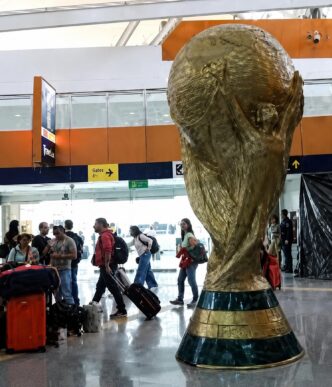 Large gold World Cup trophy sculpture in an airport terminal, with travelers and luggage around it.
