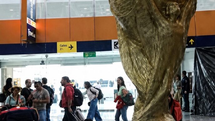 Large gold World Cup trophy sculpture in an airport terminal, with travelers and luggage around it.
