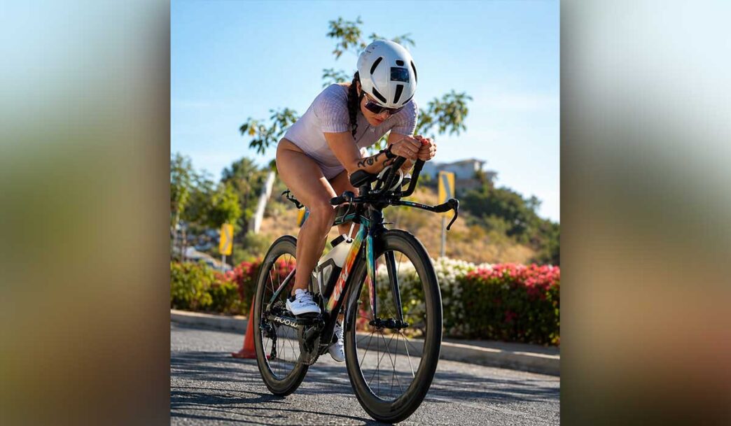 Cyclist wearing a white helmet and lavender outfit leans forward on a road bike during a sunny ride, with flowers and cones in the background.