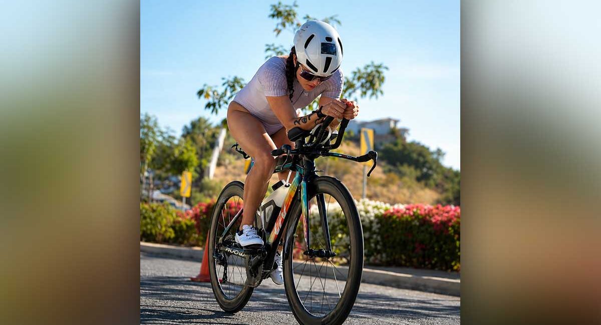 Cyclist wearing a white helmet and lavender outfit leans forward on a road bike during a sunny ride, with flowers and cones in the background.