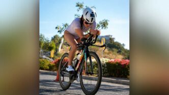 Cyclist wearing a white helmet and lavender outfit leans forward on a road bike during a sunny ride, with flowers and cones in the background.