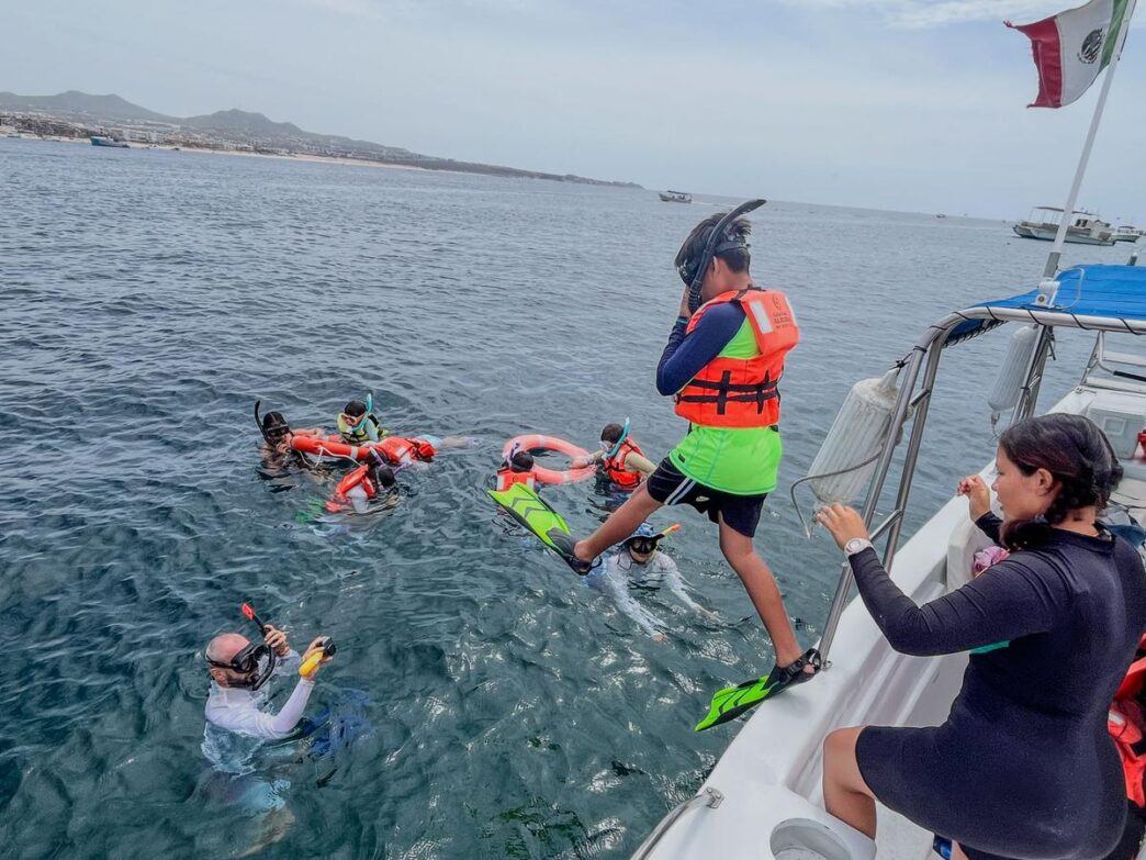 Cabo-Trek - Gringo Gazette Group of people snorkeling from a boat, wearing life jackets and fins, in clear water near shore with a Mexican flag on board.