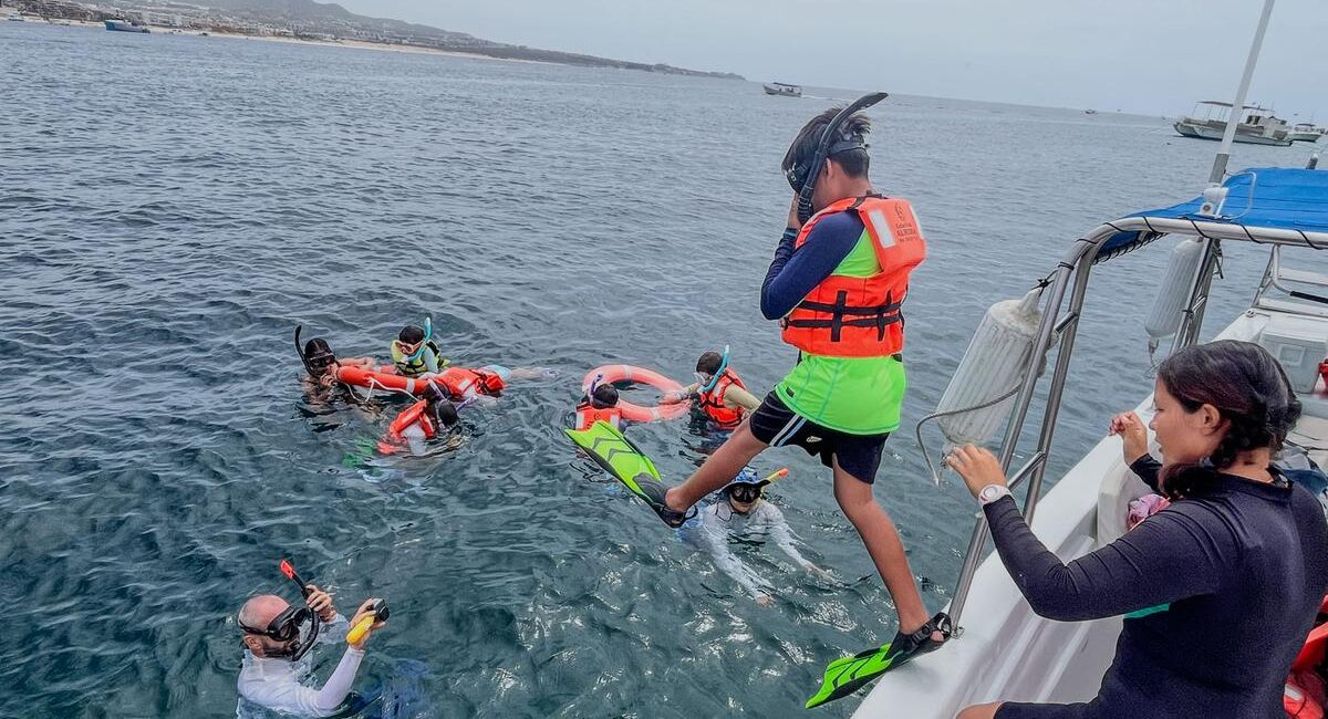 Group of people snorkeling from a boat, wearing life jackets and fins, in clear water near shore with a Mexican flag on board.
