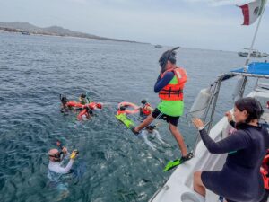 Group of people snorkeling from a boat, wearing life jackets and fins, in clear water near shore with a Mexican flag on board.