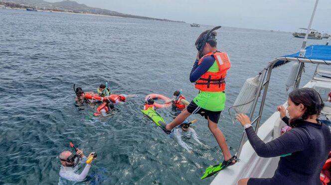 Group of people snorkeling from a boat, wearing life jackets and fins, in clear water near shore with a Mexican flag on board.