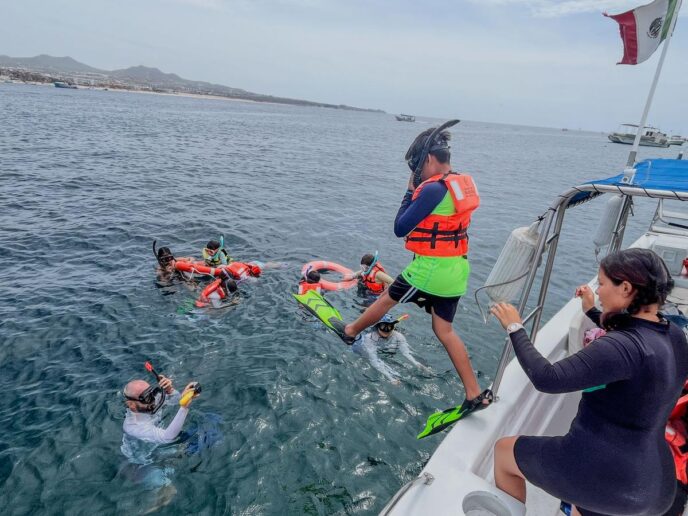 Group of people snorkeling from a boat, wearing life jackets and fins, in clear water near shore with a Mexican flag on board.
