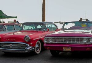 Two vintage convertibles parked side by side at an outdoor car show, a red Buick on the left and a magenta convertible on the right.