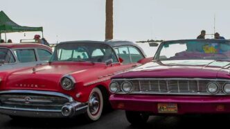 Two vintage convertibles parked side by side at an outdoor car show, a red Buick on the left and a magenta convertible on the right.