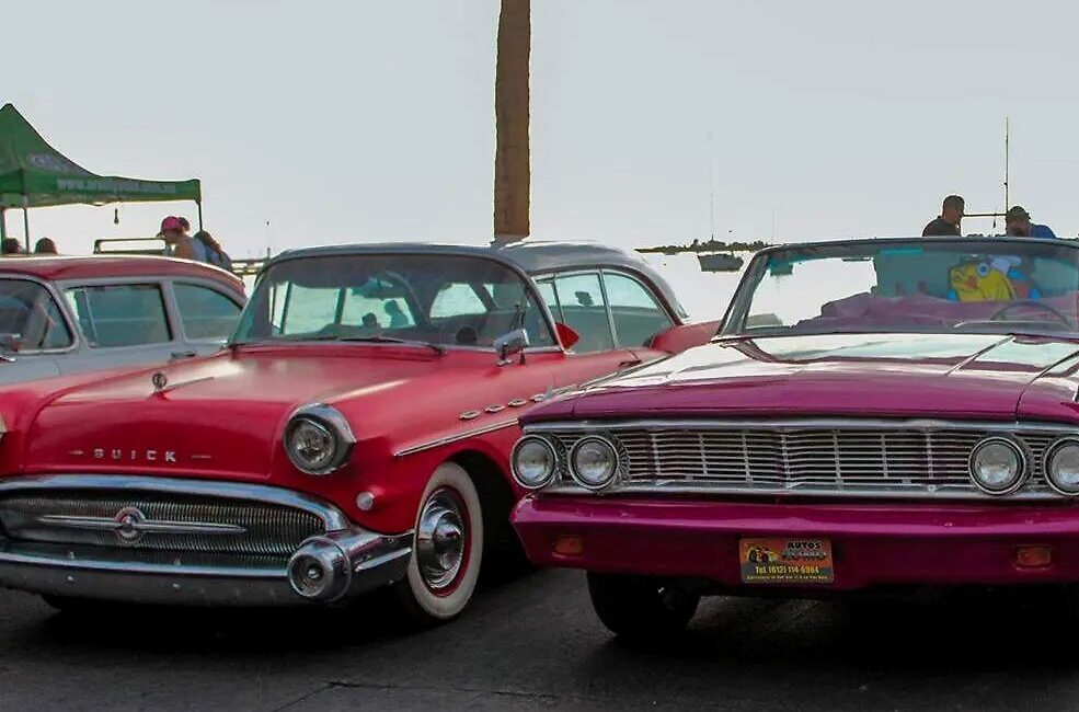 Two vintage convertibles parked side by side at an outdoor car show, a red Buick on the left and a magenta convertible on the right.