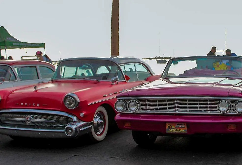 Two vintage convertibles parked side by side at an outdoor car show, a red Buick on the left and a magenta convertible on the right.
