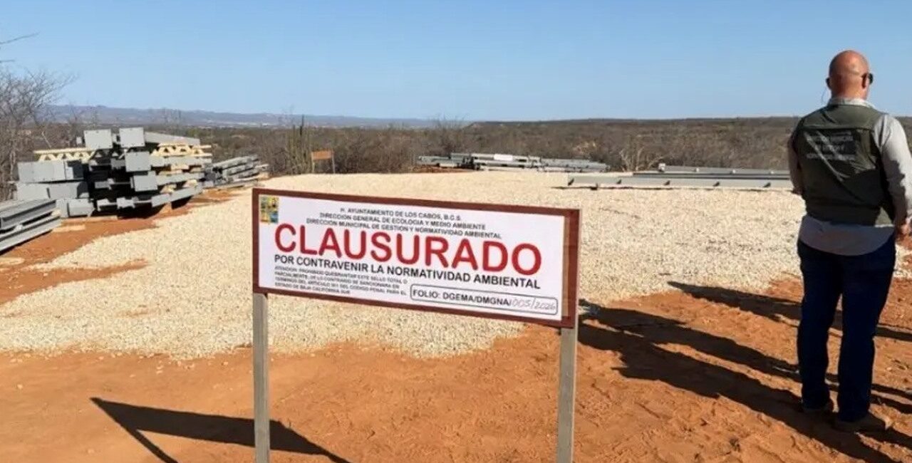 Open desert construction site with a large 'CLAUSURADO' sign; piles of metal beams and gravel on the ground, a man in a vest standing to the right.