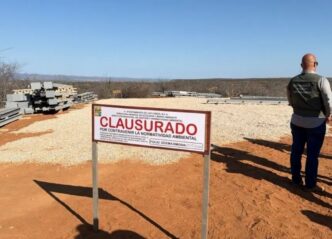 Open desert construction site with a large 'CLAUSURADO' sign; piles of metal beams and gravel on the ground, a man in a vest standing to the right.