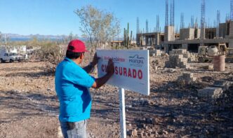 Man in a blue shirt and red cap posts a 'CERRADO' sign at an unfinished construction site under a clear blue sky.
