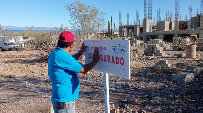 Man in a blue shirt and red cap posts a 'CERRADO' sign at an unfinished construction site under a clear blue sky.