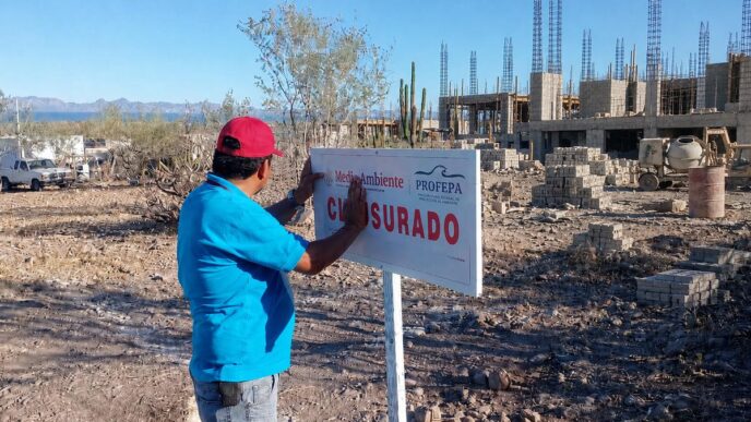 Man in a blue shirt and red cap posts a 'CERRADO' sign at an unfinished construction site under a clear blue sky.