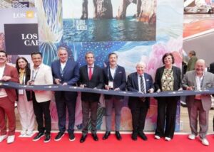 Group of dignitaries in suits and smart attire stand behind a blue ribbon on a red carpet, posing at a ribbon-cutting ceremony with a colorful Los Cabos backdrop nearby.