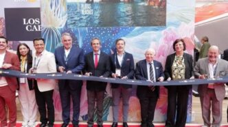 Group of dignitaries in suits and smart attire stand behind a blue ribbon on a red carpet, posing at a ribbon-cutting ceremony with a colorful Los Cabos backdrop nearby.