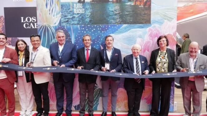 Group of dignitaries in suits and smart attire stand behind a blue ribbon on a red carpet, posing at a ribbon-cutting ceremony with a colorful Los Cabos backdrop nearby.