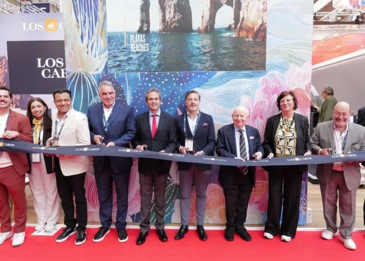 FRANKFURT - Gringo Gazette Group of dignitaries in suits and smart attire stand behind a blue ribbon on a red carpet, posing at a ribbon-cutting ceremony with a colorful Los Cabos backdrop nearby.
