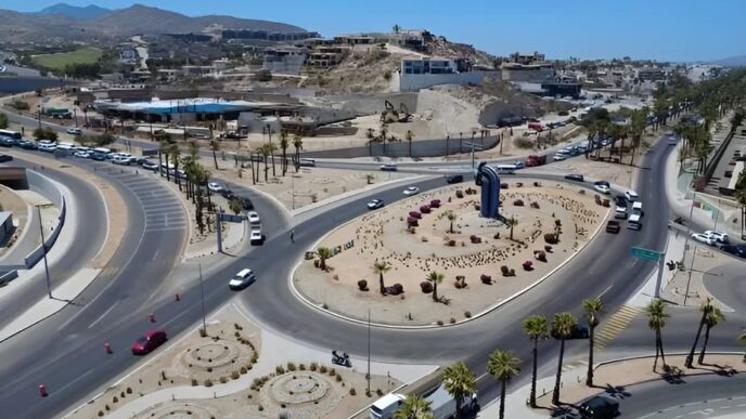 Aerial view of a large roundabout with a tall blue sculpture at the center, desert landscaping and palm trees along the roads.