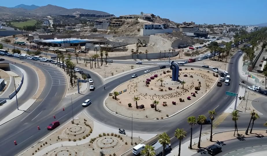 Aerial view of a large roundabout with a tall blue sculpture at the center, desert landscaping and palm trees along the roads.