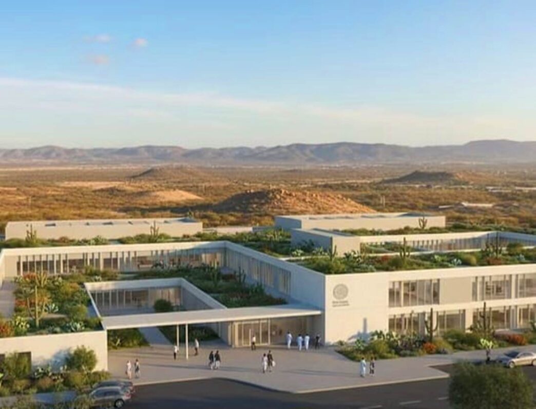 Modern white campus-style buildings with rooftop gardens in a desert landscape, hills in the distance, people walking outside.