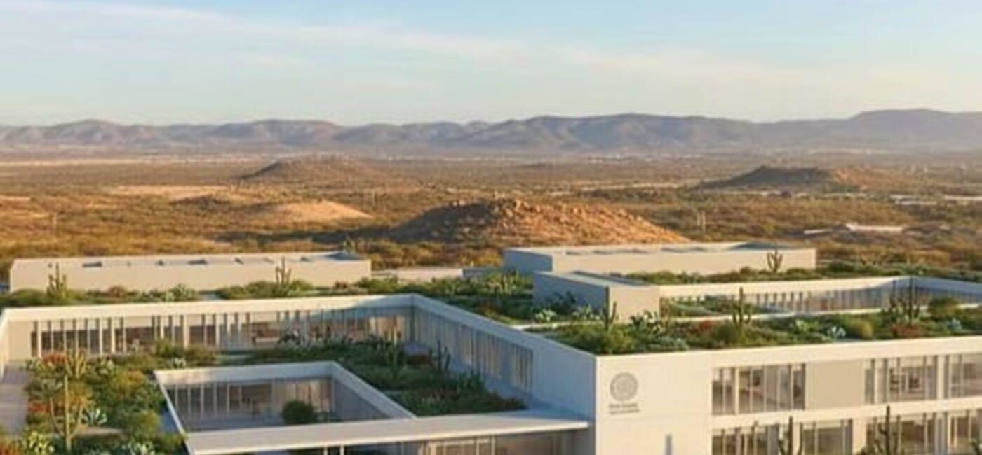 Modern white campus-style buildings with rooftop gardens in a desert landscape, hills in the distance, people walking outside.