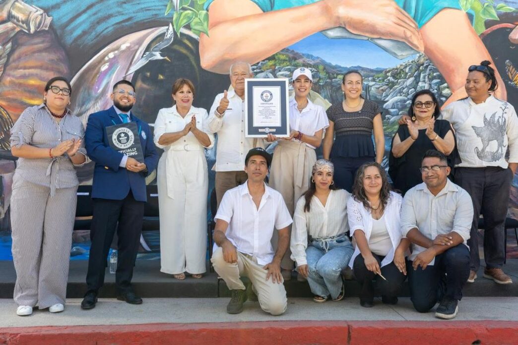 Group of 12 people posing with a framed certificate in front of a mural, some clapping and smiling.