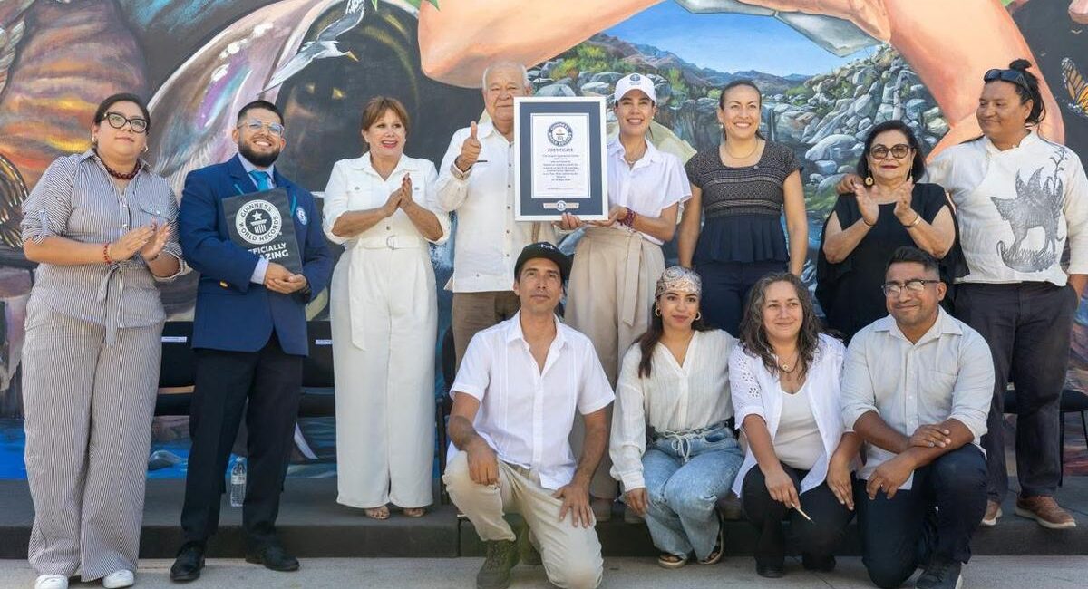 Group of 12 people posing with a framed certificate in front of a mural, some clapping and smiling.