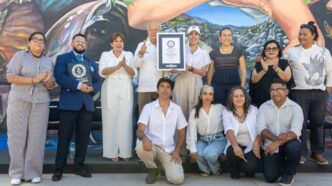 Group of 12 people posing with a framed certificate in front of a mural, some clapping and smiling.