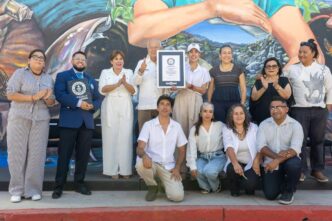 Group of 12 people posing with a framed certificate in front of a mural, some clapping and smiling.