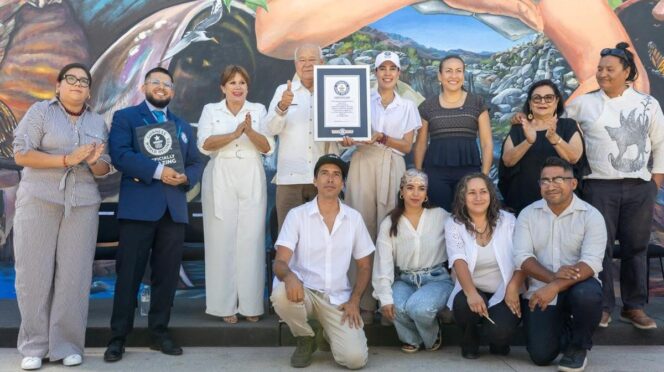 Group of 12 people posing with a framed certificate in front of a mural, some clapping and smiling.