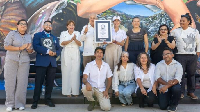 Group of 12 people posing with a framed certificate in front of a mural, some clapping and smiling.