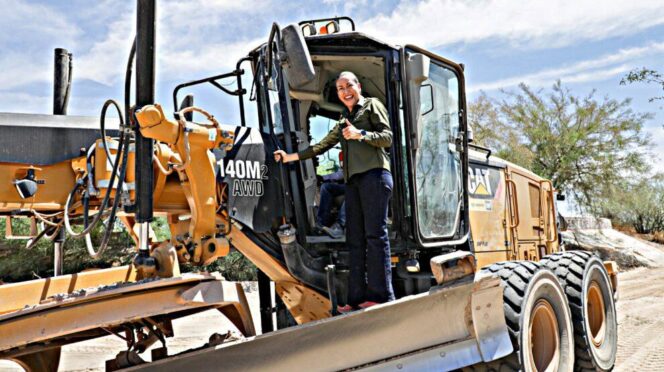 Woman in a green shirt stands in the cab of a large yellow CAT 140M AWD bulldozer, giving a thumbs-up at a sunny construction site on a dirt road.