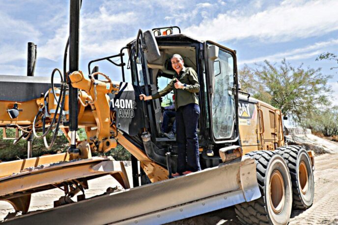 Woman in a green shirt stands in the cab of a large yellow CAT 140M AWD bulldozer, giving a thumbs-up at a sunny construction site on a dirt road.