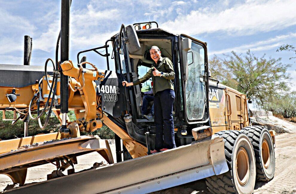 Woman in a green shirt stands in the cab of a large yellow CAT 140M AWD bulldozer, giving a thumbs-up at a sunny construction site on a dirt road.
