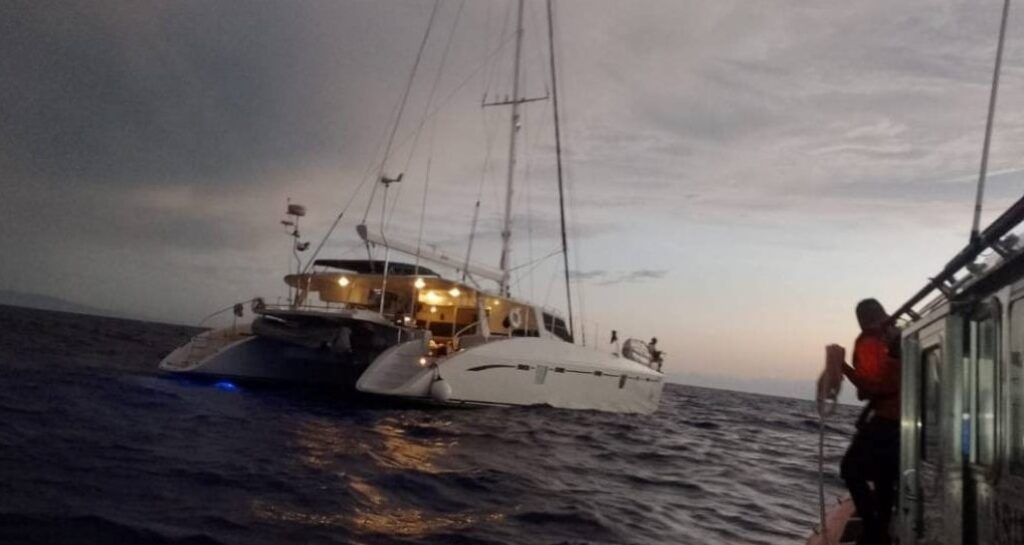 Navy2 - Gringo Gazette Two white catamaran sailboats on the open sea at dusk, with a person in an orange life jacket on a smaller boat to the right.