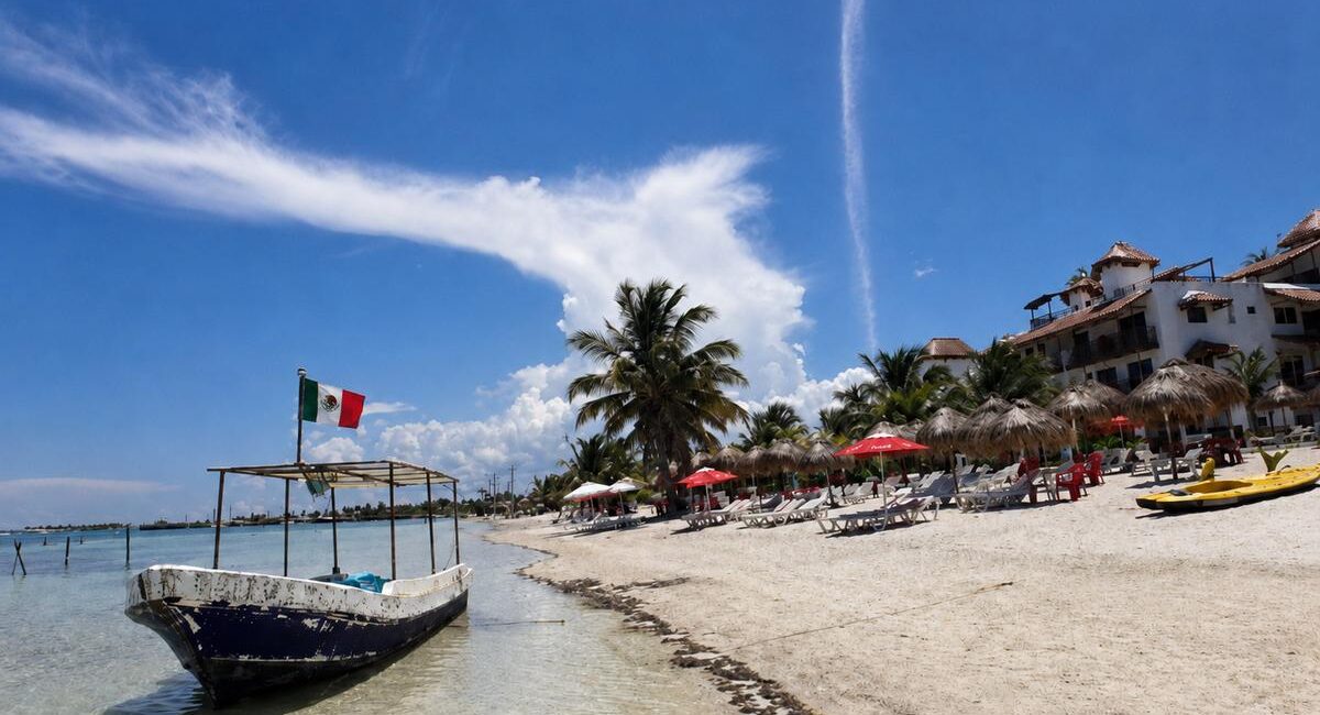 Beach scene with white sand, palm trees, wooden lounge chairs under red umbrellas, and a small boat near shore; a Mexican flag flies from a dock.