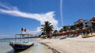 Beach scene with white sand, palm trees, wooden lounge chairs under red umbrellas, and a small boat near shore; a Mexican flag flies from a dock.