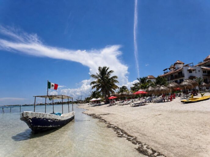 Beach scene with white sand, palm trees, wooden lounge chairs under red umbrellas, and a small boat near shore; a Mexican flag flies from a dock.