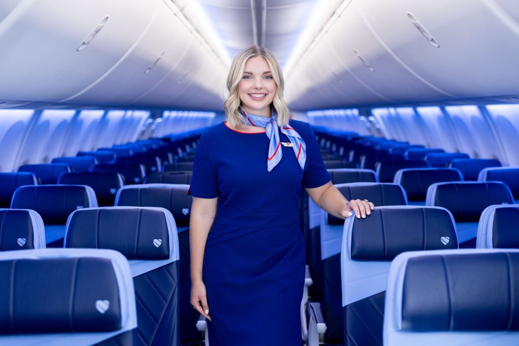 Flight attendant in a blue uniform standing in an airplane cabin with blue seats behind her in a modern jet.