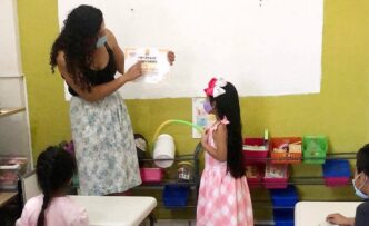 Teacher presents a diploma to a young girl in a colorful classroom while peers watch, both wearing masks.